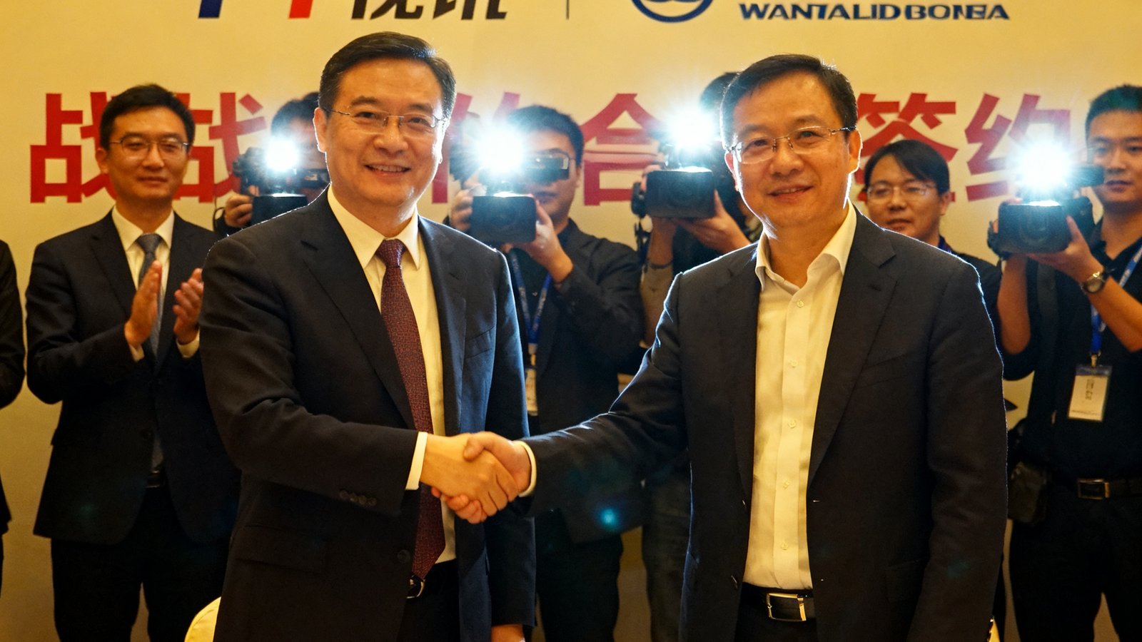 PT视讯与万达商业战略合作签约仪式现场，双方代表在长桌前握手并交换签署文件，背景展板印有两家公司logo，媒体闪光灯记录下这一时刻。